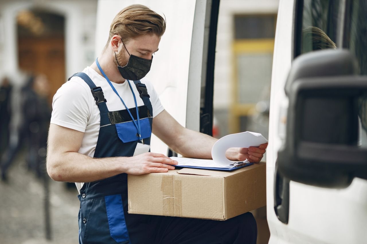 A delivery worker wearing a face mask checks a package next to a van, ensuring safety measures.